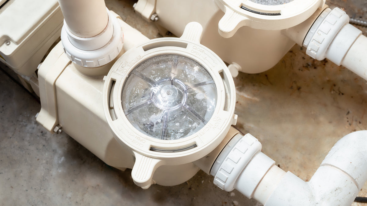 Close-up of a beige pool or spa filtration valve with a clear circular window showing internal components, connected to white PVC pipes on a concrete floor.