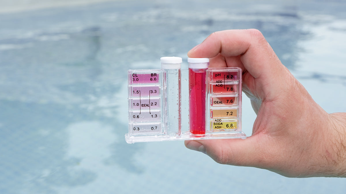 A hand holding a clear plastic pool test kit with two vials and color strips, with a blurred swimming pool in the background.